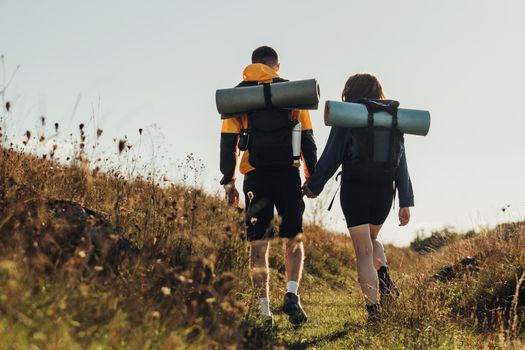 Back View of Hiking Couple, Young Travelers Man and Woman with Backpacks and Camping Mat Holding by Hands and Moving Forward on Route