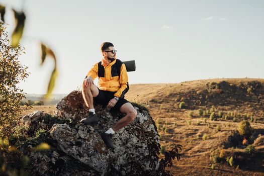 Young Traveler Man in Sunglasses Dressed in Windbreaker with Backpack, Sitting on Top of Rock During Sunset and Looking Away, Male Nomad Enjoy His Best Solo Trip