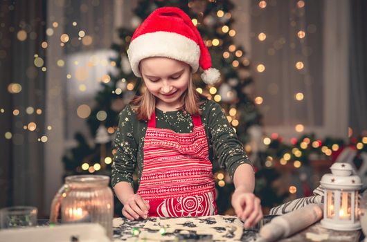 Portrait of little girl in santa hat cutting cookies on decorated christmas tree background