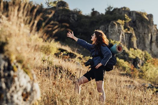 Young Traveler Woman with Backpack and Camping Mat Wearing Digital Camera on Strip and Showing Hand to Someone Outside of Shot While Hiking on Hill During Sunset