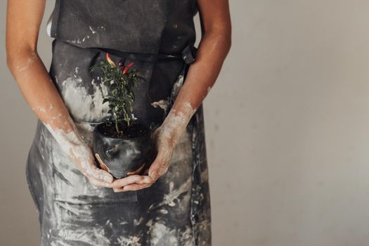 Unrecognisable Female Potter Master Dressed in Dirty Apron Holding Handmade Flowerpot with Plant in Studio