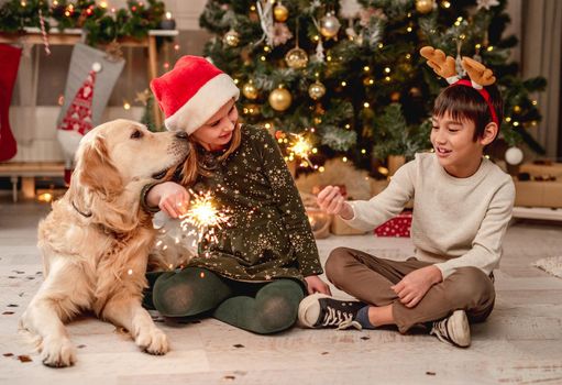 Little girl in santa hat and boy wearing reindeer horns rim holding sparklers while sitting beside golden retriever dog at home