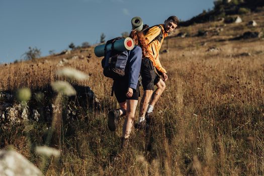 Two Travelers with Backpacks and Camping Mat on a Route, Man Helping Woman to Move Forward on Top of Hill During Sunset