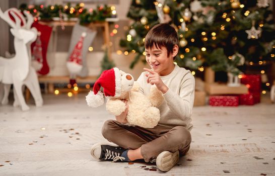 Little boy playing with teddy bear in santa hat while sitting near christmas tree at home