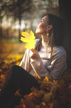 Young happy mixed race woman sitting near tree in autumn park looking at sunset.