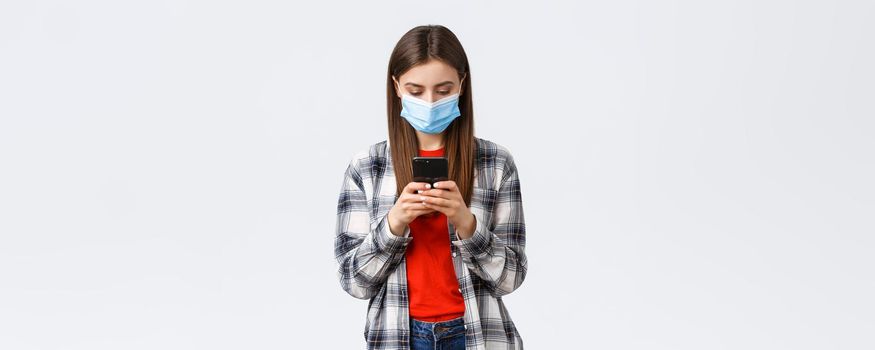 Different emotions, covid-19, social distancing and technology concept. Attractive young female in medical mask texting message, looking at mobile phone screen busy, working from home.