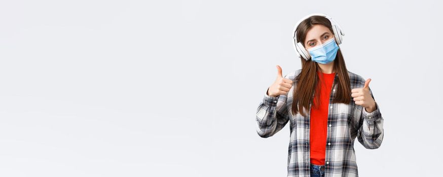 Social distancing, leisure and lifestyle on covid-19 outbreak, coronavirus concept. Pleased good-looking woman in medical mask, listening music headphones, show thumb-up good headsets.