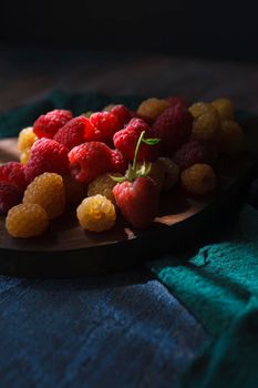 Red and yellow raspberry on wooden board on blue and green background, close up, selective focus low key.