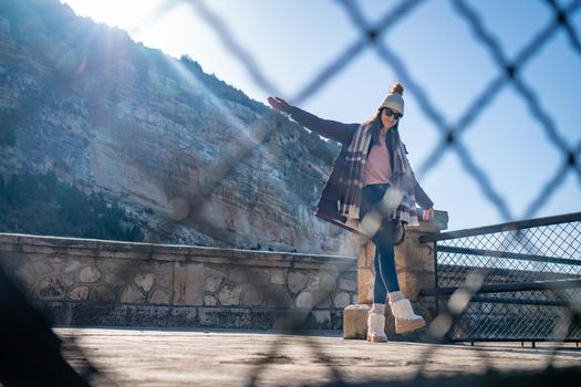 Young woman standing with outstretched hands wearing coat and hat behind a fence outdoors