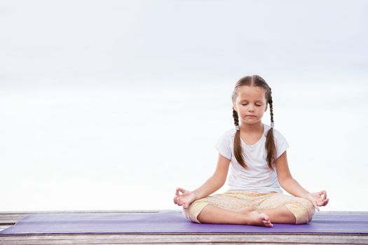 Child doing meditating exercise on wooden platform sea shore outdoors. Healthy lifestyle. Yoga girl