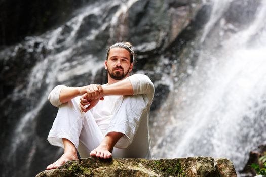 Man sitting in meditation yoga on rock at waterfall in tropical rainforest