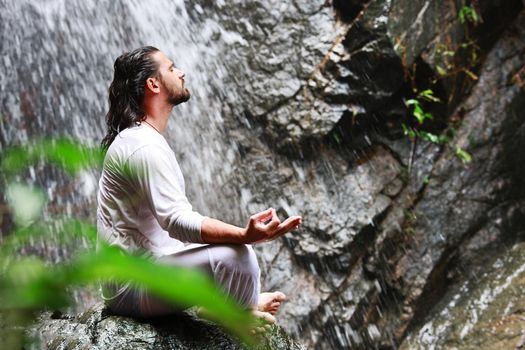 Man sitting in meditation yoga on rock at waterfall in tropical rainforest