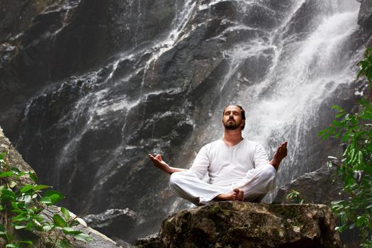Man sitting in meditation yoga on rock at waterfall in tropical rainforest