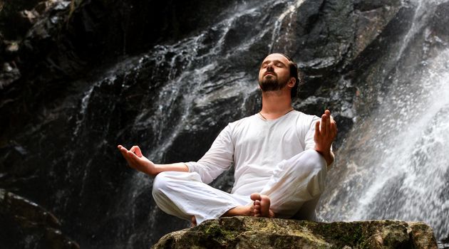 Man sitting in meditation yoga on rock at waterfall in tropical rainforest