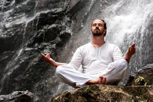 Man sitting in meditation yoga on rock at waterfall in tropical rainforest