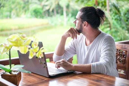 Freelance work on laptop. Casual dressed man sitting at wooden desk inside garden working on computer