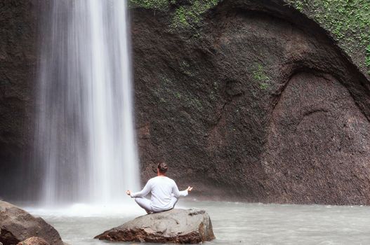 Man sitting in meditation yoga on rock at waterfall in tropical. Tibumana Waterfall