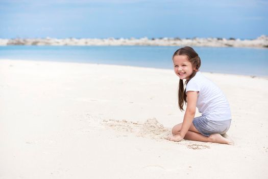 Happy child on the beach. Paradise holiday concept, girl in preschool age standing on sandy beach with blue shallow water and clean sky on background