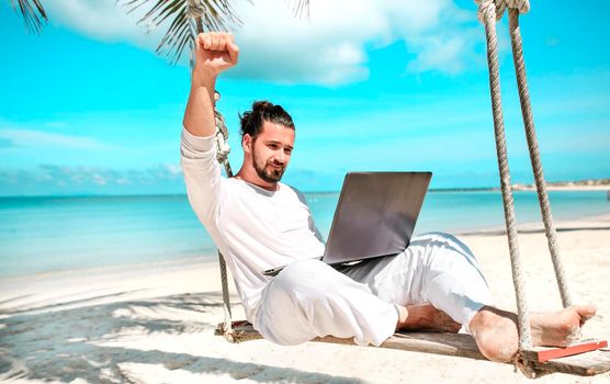 Man working with a laptop, on a hammock in the beach. Concept of digital nomad, remote worker, independent location entrepreneur. Show win