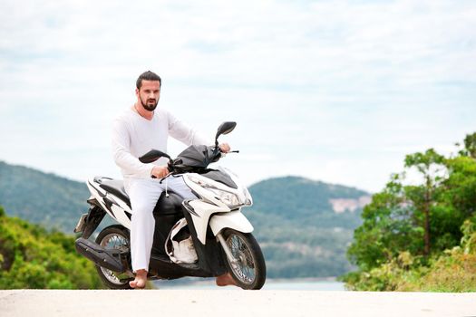 Brutal biker with beard in white sitting on motorbike. Sunny day in the mountains.