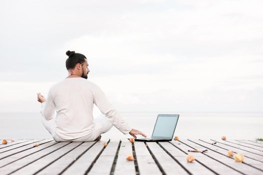 Caucasian man in white clothes meditating yoga with laptop on wooden pier platform