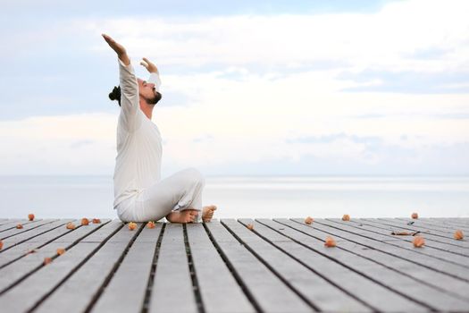 Caucasian man in white clothes meditating yoga with laptop on wooden pier