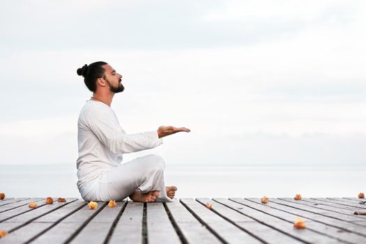 Caucasian man in white clothes meditating yoga on wooden pier platform