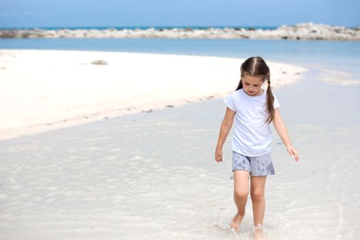 Happy child on the beach. Paradise holiday concept, girl in preschool age standing on sandy beach with blue shallow water and clean sky on background