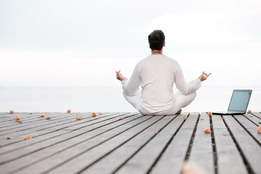 Caucasian man in white clothes meditating yoga with laptop on wooden pier platform