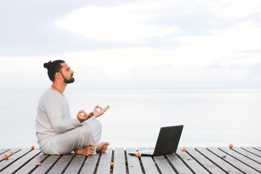 Caucasian man in white clothes meditating yoga with laptop on wooden pier