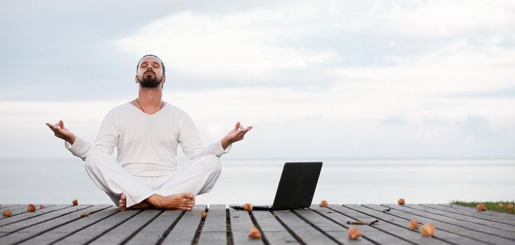 Caucasian man in white clothes meditating yoga with laptop on wooden pier