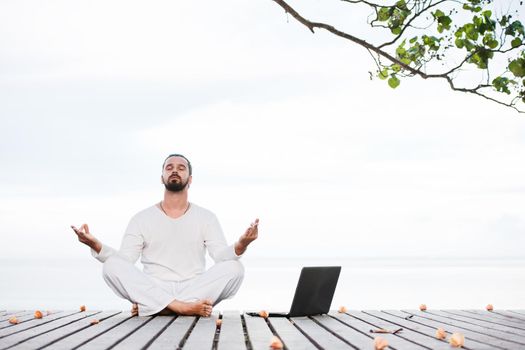 Caucasian man in white clothes meditating yoga with laptop on wooden pier