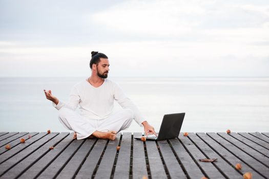 Caucasian man in white clothes meditating yoga with laptop on wooden pier