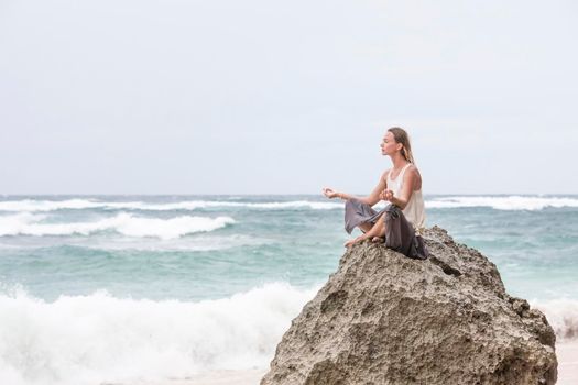 Beautiful girl clothing in white sit at the seaside on the rock and meditating in yoga woman pose