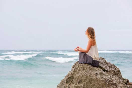 Beautiful girl clothing in white sit at the seaside on the rock and meditating in yoga woman pose