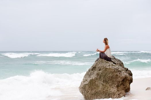 Beautiful girl clothing in white sit at the seaside on the rock and meditating in yoga woman pose