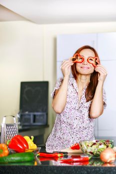 Portrait of beautiful woman in apron covering her eyes with pepper and smiling while standing in the kitchen