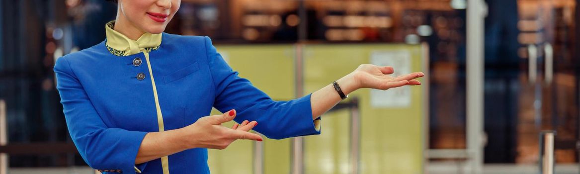Cheerful female flight attendant in air hostess uniform gesturing towards something and smiling while standing in airport terminal