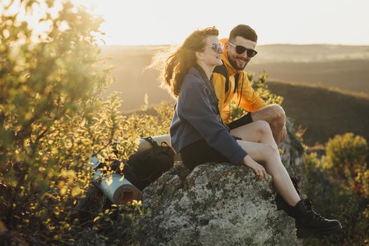 Happy Traveler Couple Man and Woman Sitting on Edge of Rock During Sunset and Smiling, Wind Blowing Woman's Hair