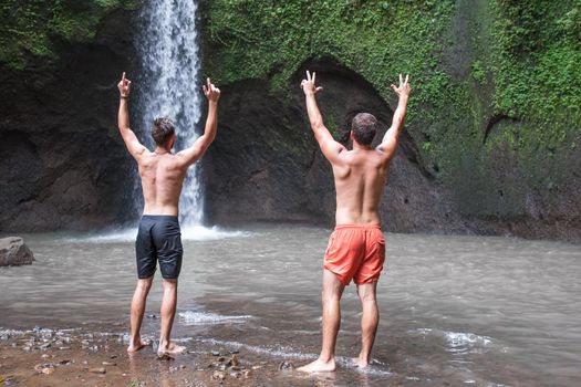 Two men with hands up. Picture of nature and beautiful Tibumana waterfall in bali