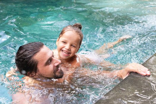 Happy family, active father with little child, adorable toddler girl, having fun together in outdoors swimming pool in water park during sunny summer sea vacation in tropical resort.