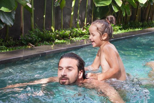 Happy family, active father with little child, adorable toddler girl, having fun together in outdoors swimming pool in water park during sunny summer sea vacation in tropical resort.