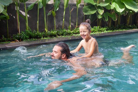 Happy family, active father with little child, adorable toddler girl, having fun together in outdoors swimming pool in water park during sunny summer sea vacation in tropical resort.