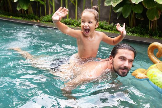 Happy family, active father with little child, adorable toddler girl, having fun together in outdoors swimming pool in water park during sunny summer sea vacation in tropical resort.