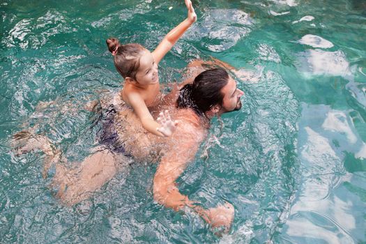Happy family, active father with little child, adorable toddler girl, having fun together in outdoors swimming pool in water park during sunny summer sea vacation in tropical resort.