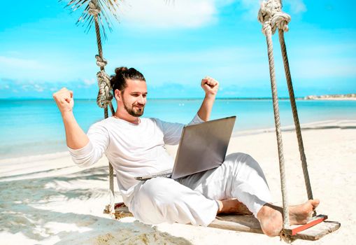 Man working with a laptop, on a hammock in the beach. Concept of digital nomad, remote worker, independent location entrepreneur. Show win