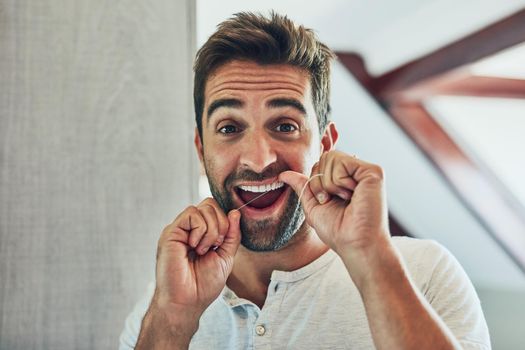 Portrait of a cheerful young man flossing his teeth while looking at his reflection in the mirror at home.