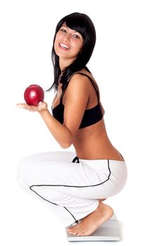 Happy young athlete woman standing on weighting scales and holding red apple isolated on white background