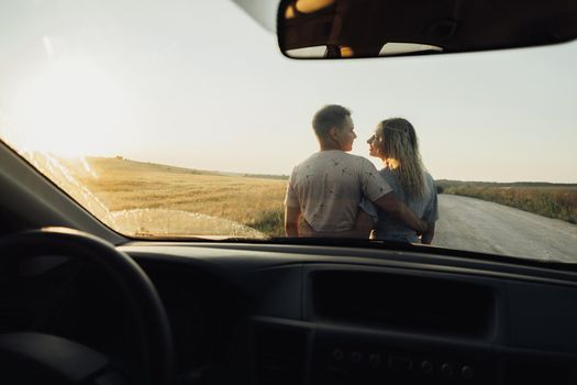 Back View Through Front Car Window on Young Couple Hugging Near the Hood, Woman and Man Enjoying Road Trip at Sunset