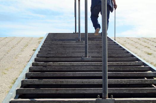 The legs of an elderly man with cane. He climbs a public staircase on a dike.